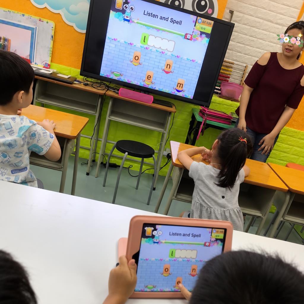 A tutor leads a 'Listen and Spell' activity on the interactive whiteboard while students follow along on a tablet